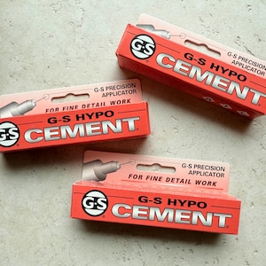 May include: Three red and white G-S Hypo Cement boxes. Each box features the text "G-S HYPO CEMENT" in large, silver letters, and "G-S Precision Applicator" above. The boxes are designed for fine detail work.