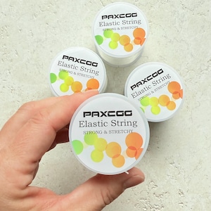 May include: Four spools of PAXCOG Elastic String are displayed. Each spool has a white label with the brand name and product description. The labels feature a colorful design with green, yellow, and orange circles. The text on the label reads "STRONG & STRETCHY".