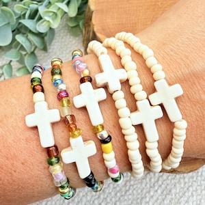 May include: A collection of beaded bracelets featuring white cross charms. Some bracelets have colorful beads, while others have cream-colored beads. The bracelets are displayed on a person's wrist, showcasing the jewelry's design and style.
