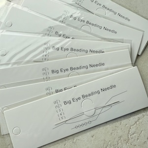 May include: A stack of white cardstock packages, each containing a big eye beading needle. The packages are labeled with the text "Big Eye Beading Needle" and include needle size measurements in inches. A diagram of the needle is also shown.