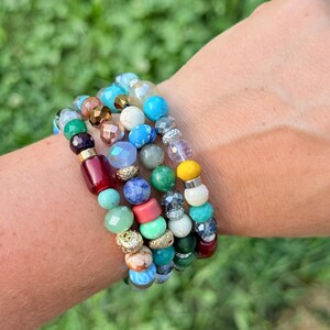 Puede incluir: Una colecci&oacute;n de pulseras de cuentas en varios colores, incluyendo turquesa, rojo, verde y dorado. Las pulseras presentan una mezcla de cuentas redondas y facetadas, creando un aspecto en capas. Las pulseras est&aacute;n apiladas en una mu&ntilde;eca sobre un fondo verde borroso.