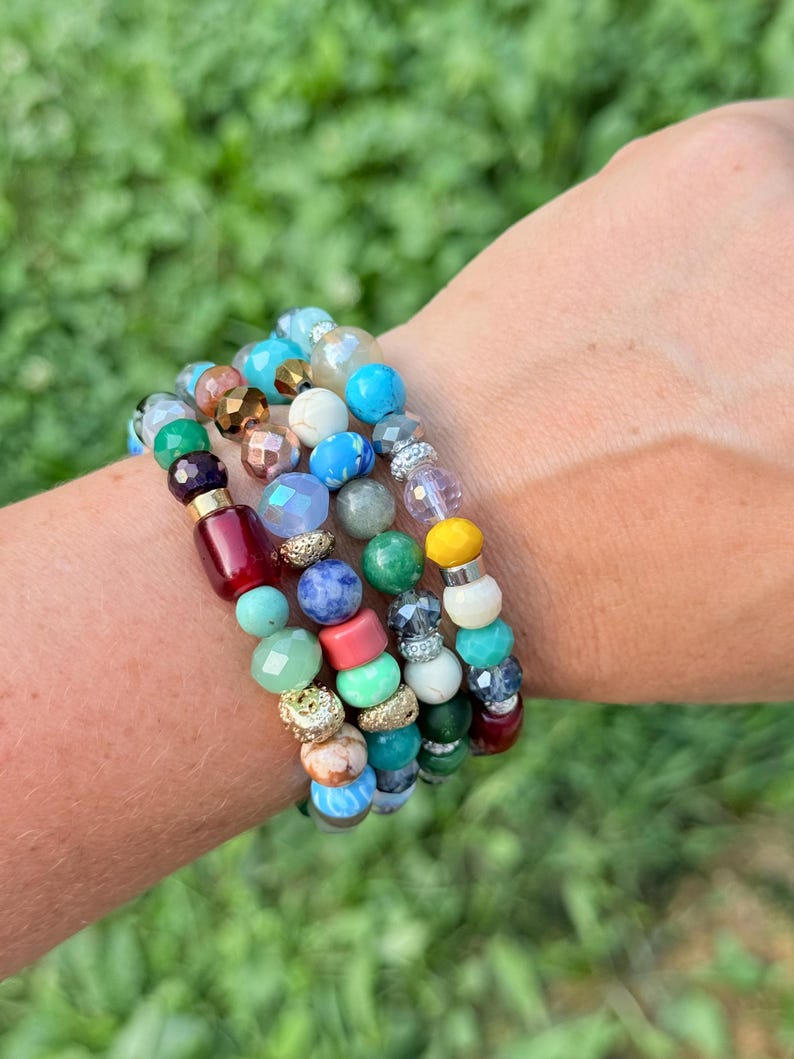 Puede incluir: Una colecci&oacute;n de pulseras de cuentas en varios colores, incluyendo turquesa, rojo, verde y dorado. Las pulseras presentan una mezcla de cuentas redondas y facetadas, creando un aspecto en capas. Las pulseras est&aacute;n apiladas en una mu&ntilde;eca sobre un fondo verde borroso.