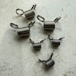 May include: A collection of six small, silver-colored metal coil springs with attached loops. The springs are tightly wound and appear to be made of a shiny, durable material. They are arranged on a light-colored, speckled surface.