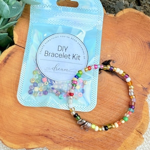 May include: A DIY bracelet kit with a completed bracelet. The kit includes a clear plastic bag with colorful beads and the text "DIY Bracelet Kit". The bracelet is made of various colorful beads and a silver charm. The kit and bracelet are on a wooden surface.