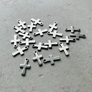 May include: A collection of small, silver-colored cross charms scattered on a light gray surface. Each charm has a loop at the top for attaching to jewelry or other crafts. The crosses are simple in design and appear to be made of metal.