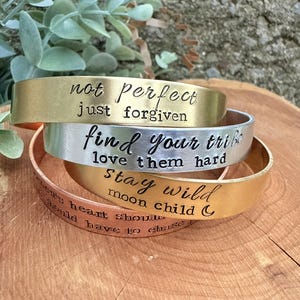 May include: Four metal cuff bracelets stacked on a wooden surface. The bracelets are gold, silver, and copper-toned, each engraved with inspirational phrases like "not perfect just forgiven" and "find your tribe love them hard."