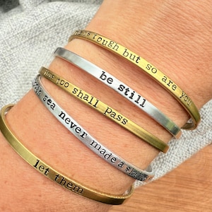 May include: A collection of five metal bangles in gold and silver tones. Each bangle features an engraved inspirational quote, including "This too shall pass" and "Be still". The bangles are stacked on a wrist.
