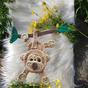 May include: A crocheted monkey toy in shades of tan and brown, hanging from a brown branch with green leaves. The monkey has a smiling face and is set against a white, fluffy background with yellow flowers.
