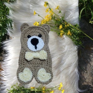 May include: A crocheted brown bear with a yellow bow tie and yellow heart-shaped feet. The bear is sitting on a white fluffy surface.