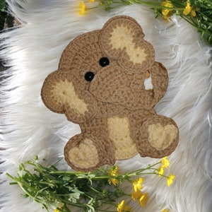 May include: A crocheted brown and cream elephant toy with a heart-shaped design on the feet. The toy is sitting on a white fluffy surface.