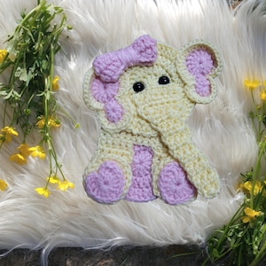 May include: A crocheted elephant figure crafted from pale yellow yarn with light pink accents on the ears, feet, and center body. The elephant features a prominent trunk, two black bead eyes, and a textured pink bow attached to the top of its head.