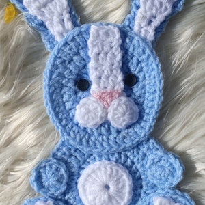 May include: A crocheted blue bunny with white accents. The bunny has a white nose, white ears, and white feet. The bunny is sitting on a white fluffy surface.
