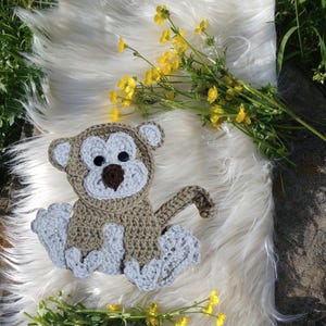 May include: A handmade crochet monkey in shades of brown, gray, and white, with black button eyes and a brown nose. The monkey is sitting on a white, fluffy surface, with yellow flowers and green foliage in the background.