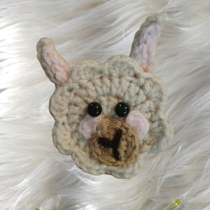 May include: A crocheted llama with a brown nose and black eyes. The llama is white with pink ears and cheeks.