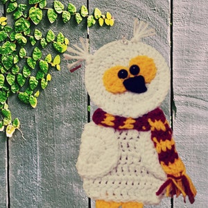 May include: White crocheted owl with yellow eyes and a red and yellow scarf. The owl is hanging on a wooden wall with green vines.