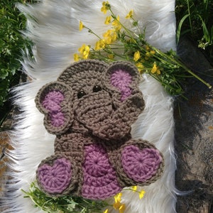 May include: A crocheted elephant toy with brown and purple yarn. The elephant has large ears and a trunk. The toy is sitting on a white fluffy surface.