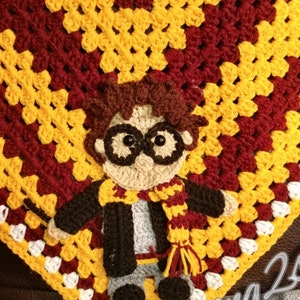 May include: A handmade crochet blanket featuring a character design with round glasses, a scarf, and a wand. The blanket is primarily burgundy, yellow, and white, with a character design in the center. The blanket is made with a granny square pattern.