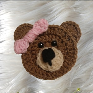 May include: A crocheted brown bear face with a pink bow. The bear has a tan muzzle, black eyes, and a black nose. The handmade item is likely a decorative applique or embellishment for crafts.