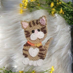 May include: A crocheted brown and white cat with a pink nose and orange collar. The cat is sitting on a white fluffy surface.