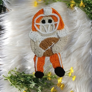 May include: A crocheted football player holding a football. The player is wearing an orange and white uniform with a white helmet. The football is brown and white.