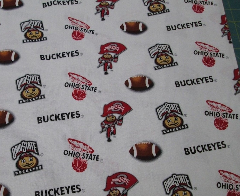 May include: A white fabric featuring a repeating pattern of Ohio State Buckeyes logos, brown footballs, red basketballs in hoops, and a mascot character holding an Ohio State flag. The text BUCKEYES and OHIO STATE is printed throughout the design.