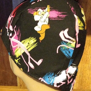 May include: Black welding cap with a colorful pattern of pink flamingos and a cartoon character in a white coat. The pattern is on a black background with colorful brushstrokes.