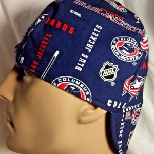 May include: Blue and red fabric welding cap with the Columbus Blue Jackets NHL logo repeated throughout the fabric. The cap has a flap that can be tied at the back of the head.