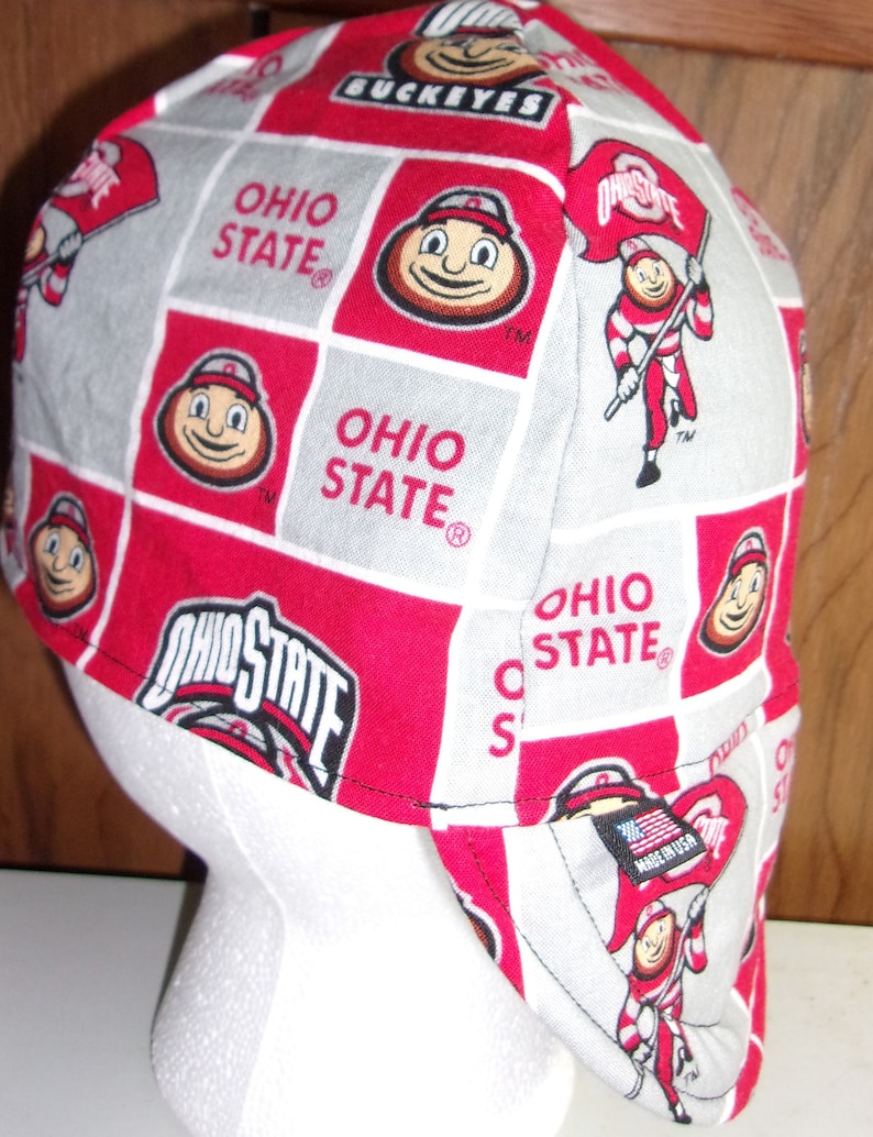 May include: A red and gray fabric cap featuring a repeating pattern of the Ohio State University logo, the Brutus Buckeye mascot, and the text Ohio State. The cap includes a small black tag on the side with an American flag and the text Made in USA.