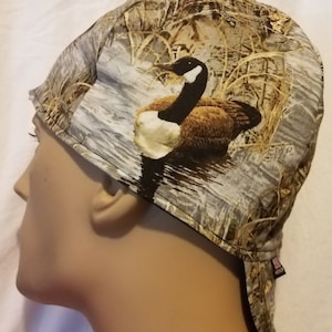 May include: A brown, gray, and white camouflage welding cap with a goose design. The cap has a flap that can be worn down to protect the neck.