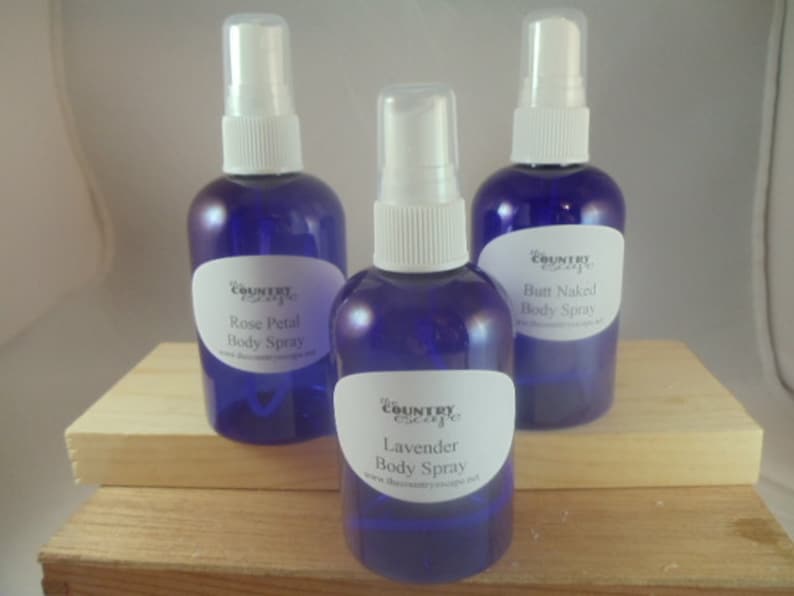 Puede incluir: Tres botellas de vidrio azul con tapas de pulverizaci&oacute;n blancas y etiquetas blancas. Las etiquetas tienen texto negro que dice "The Country Escape" y el aroma de cada spray corporal: "Rose Petal Body Spray", "Butt Naked Body Spray" y "Lavender Body Spray".