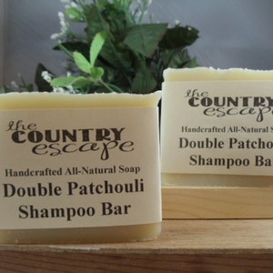 May include: Two rectangular shampoo bars with cream-colored tops and labels. The labels read "the COUNTRY escape Handcrafted All-Natural Soap Double Patchouli Shampoo Bar". The bars are on a wooden surface, with greenery in the background.