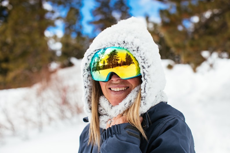 Puede incluir: Una mujer con una chaqueta de esqu&iacute; azul con capucha forrada de piel blanca y gafas de esqu&iacute; amarillas y azules. Las gafas reflejan una escena de bosque nevado.