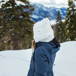 Puede incluir: Una persona con un gorro de punto blanco y una chaqueta azul se encuentra en un paisaje nevado. La persona est&aacute; mirando hacia una cordillera en la distancia.