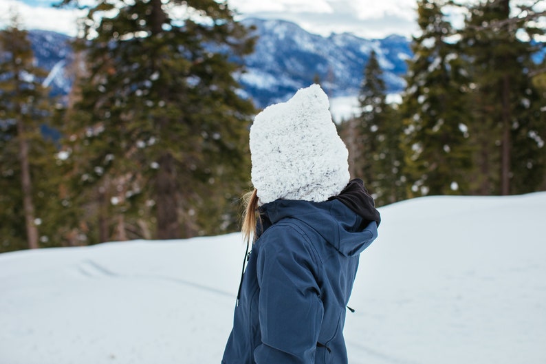 Puede incluir: Una persona con un gorro de punto blanco y una chaqueta azul se encuentra en un paisaje nevado. La persona est&aacute; mirando hacia una cordillera en la distancia.