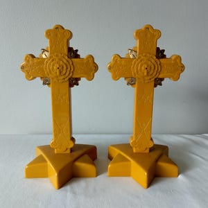May include: Two identical mustard yellow plastic crosses standing on star-shaped bases. Each cross features embossed geometric symbols and a central circular emblem with metallic gold accents. The design includes intricate engravings on the vertical and horizontal arms of the cross structure.