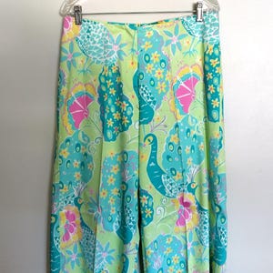 May include: High-waisted trousers featuring a vibrant, multi-coloured print of stylised peacocks and floral patterns in shades of lime green, turquoise, bright pink, and yellow. The trousers have a straight-leg cut and are displayed on a clear hanger.
