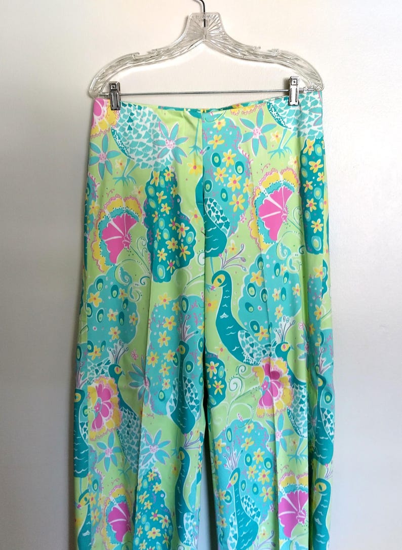 May include: High-waisted trousers featuring a vibrant, multi-coloured print of stylised peacocks and floral patterns in shades of lime green, turquoise, bright pink, and yellow. The trousers have a straight-leg cut and are displayed on a clear hanger.
