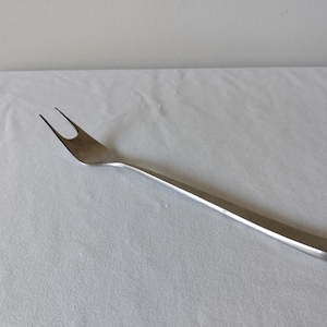 May include: A stainless steel carving fork with two tines. The fork has a long, flat handle and is designed for serving meats. The fork is shown on a white surface.
