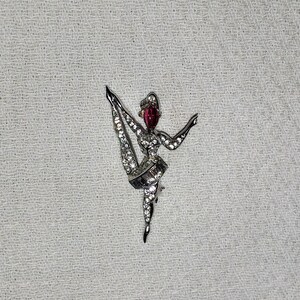 May include: A silver-toned ballerina brooch with clear and red rhinestones. The brooch depicts a ballerina in a split, with a red gemstone head and clear rhinestone accents on the body and tutu. The brooch is set against a textured white background.