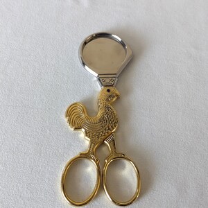 May include: Gold-toned rooster-shaped scissors with a silver-colored ring at the top. The scissors feature a detailed rooster design with a textured body and a small black eye. The blades are oval-shaped.