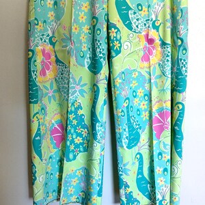 May include: A pair of wide-leg trousers featuring a vibrant, repeating pattern of teal peacocks, bright yellow flowers, and magenta hibiscus blossoms set against a light lime green background. The trousers have a straight-leg cut and a pressed crease.