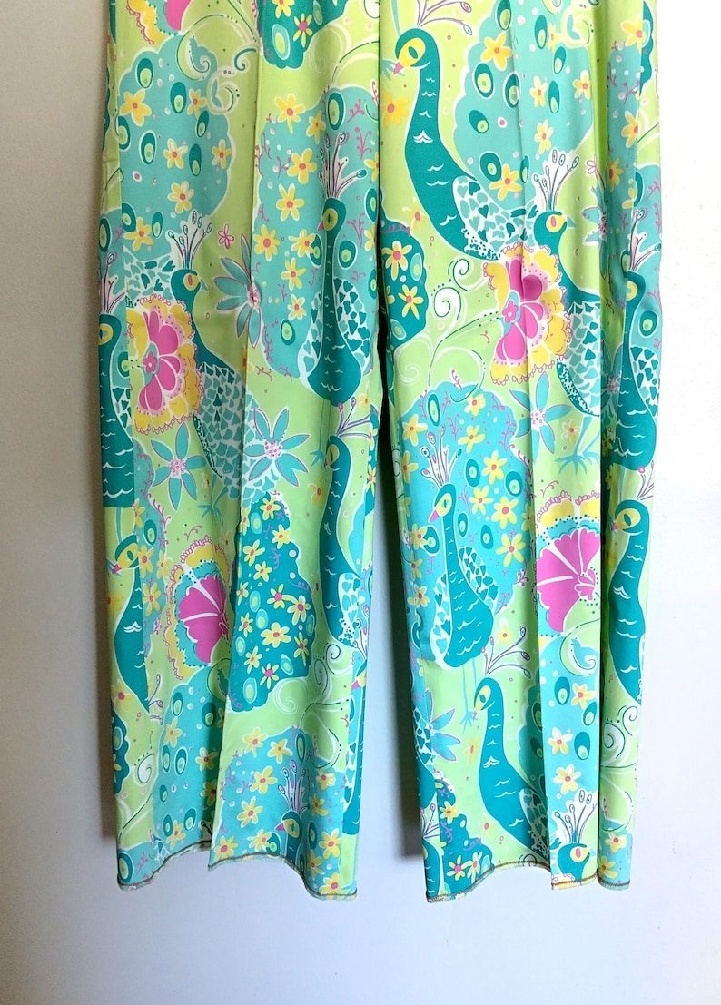 May include: A pair of wide-leg trousers featuring a vibrant, repeating pattern of teal peacocks, bright yellow flowers, and magenta hibiscus blossoms set against a light lime green background. The trousers have a straight-leg cut and a pressed crease.