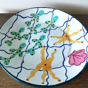 May include: A white ceramic plate with a blue rim and a whimsical underwater design. The plate features turquoise fish with black spots, yellow starfish, a pink seashell, and blue wavy lines. The design is hand-painted.