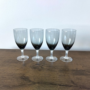 May include: Four clear glass wine glasses with a dark gray tint at the bottom. The glasses are arranged in a row on a wooden surface.