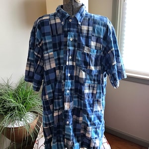 May include: A short-sleeve button-down shirt featuring a patchwork design composed of various blue, white, and navy plaid fabric squares. The shirt includes a standard collar, a front button placket with white buttons, and a single chest pocket.