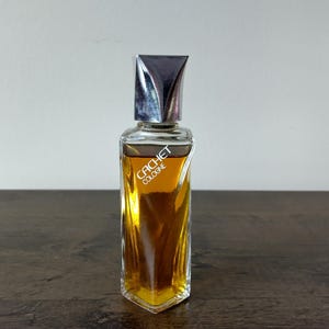 May include: A vintage bottle of "Cachet Cologne" with a silver cap. The clear glass bottle contains a golden-brown liquid. The bottle has a rectangular shape with a slightly twisted design. The text "Cachet Cologne" is printed on the front.