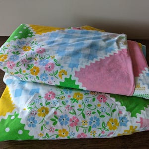 May include: Two folded vintage pillowcases. One pillowcase has a green, yellow, and white scalloped edge with a floral pattern. The other pillowcase has a pink, blue, and white gingham pattern.