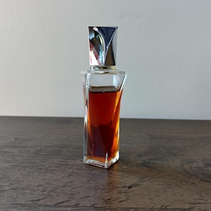 May include: A small, clear glass perfume bottle with a silver cap. The bottle has a unique, twisted shape and is filled with a dark amber liquid. The cap is a shiny silver color. The bottle is sitting on a dark wooden surface.