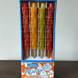 May include: A box of four colorful tiki-shaped mini torches. The torches are red, orange, green, and pink, and are on wooden sticks. The box has the text "BOSTON WAREHOUSE" and "BALI HAI mini torches".