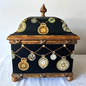 May include: A black decorative box with a hinged lid, adorned with gold-colored clock pendants hanging from chains. The box has a gold-colored trim and ornate feet, creating a vintage aesthetic. The top of the box has a decorative finial.